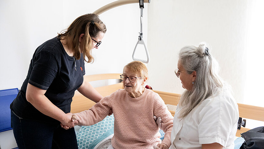 Une aide soignante et une infirmière aident une patiente à se lever de son lit médicalisé.